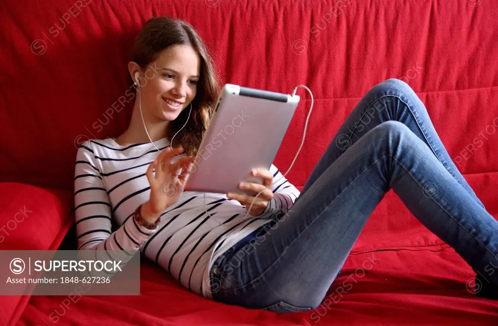 Girl lying on a sofa listening to music holding a iPad, tablet computer with wireless internet access
