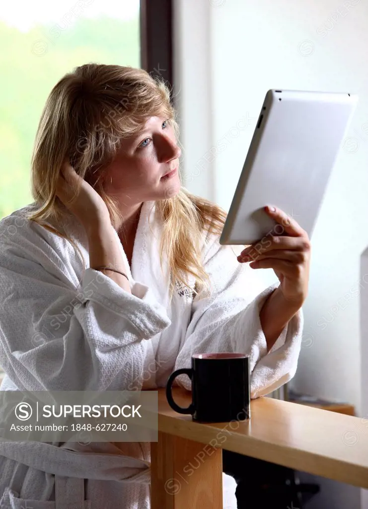 Young woman wearing a dressing gown drinking a cup of coffee in the morning, sitting in the kitchen reading an online newspaper on an iPad, a tablet c...