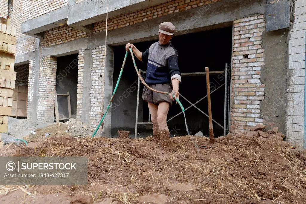 Loam rendering, building site, Muslim Uighur worker, standing amidst a mud and loam rendering mixture and is trampling down the mixture with water, in...