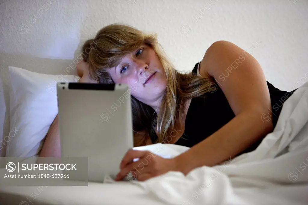 Young woman lying in bed surfing the internet on an iPad, tablet computer with wireless internet access