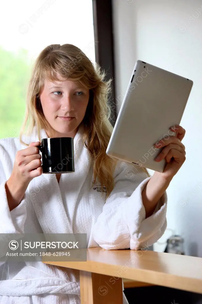 Young woman wearing a dressing gown drinking a cup of coffee in the morning, sitting in the kitchen reading an online newspaper on an iPad, a tablet c...