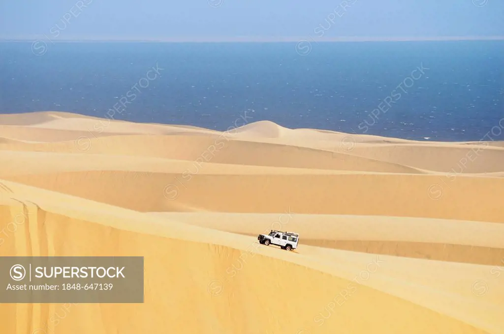 Landrover Defender off-road vehicle driving on a huge dune, Atlantic Ocean at the back, Namib Naukluft National Park, part of the Namibian Skeleton Co...