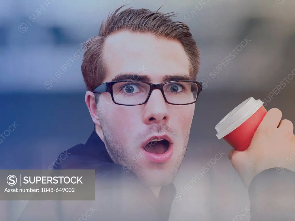 Young businessman wearing glasses, holding a cup of coffee, surprised face
