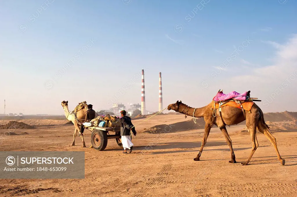 Two Indian men passing an industrial plant with two huge chimneys with their camels and a camel cart, Thar Desert, Rajasthan, India, Asia