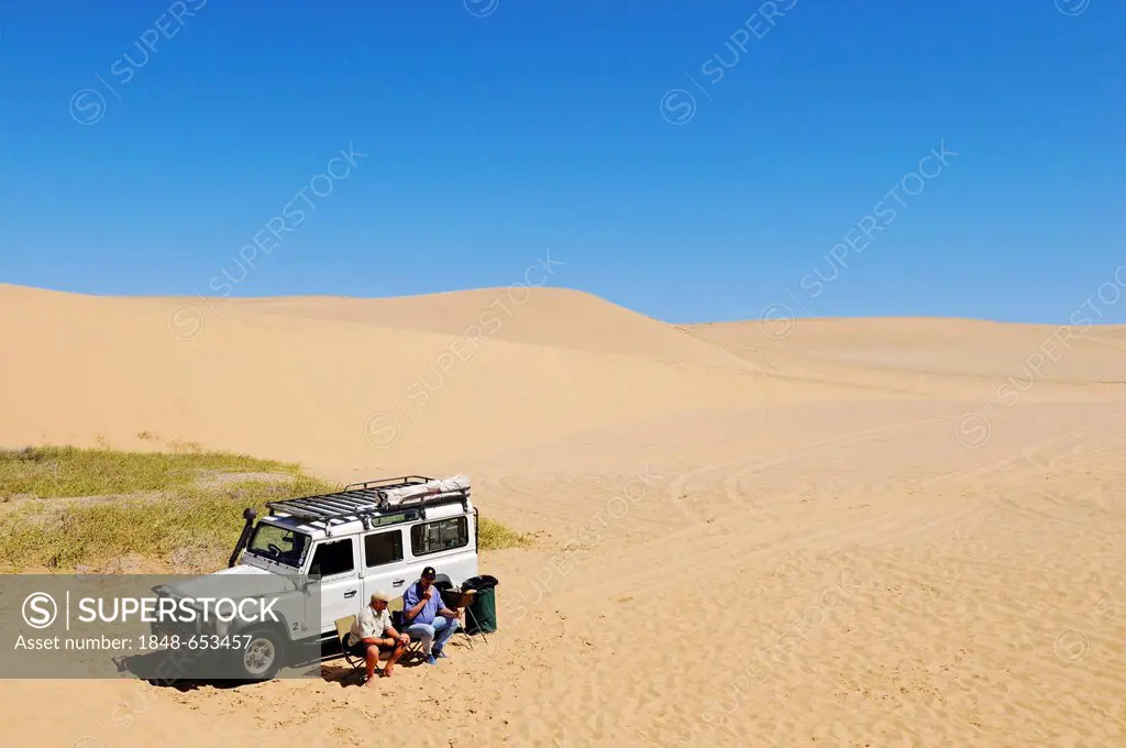 Two men sitting in front of a Landrover Defender off-road vehicle in the dunes of the Namib Naukluft National Park, part of the Namibian Skeleton Coas...