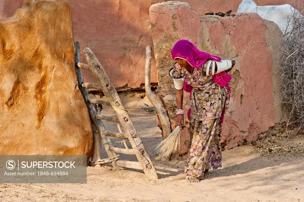 Young Indian woman wearing a sari sweeping the floor in front of her yard gate with a straw broom, Thar Desert, Rajasthan, India, Asia