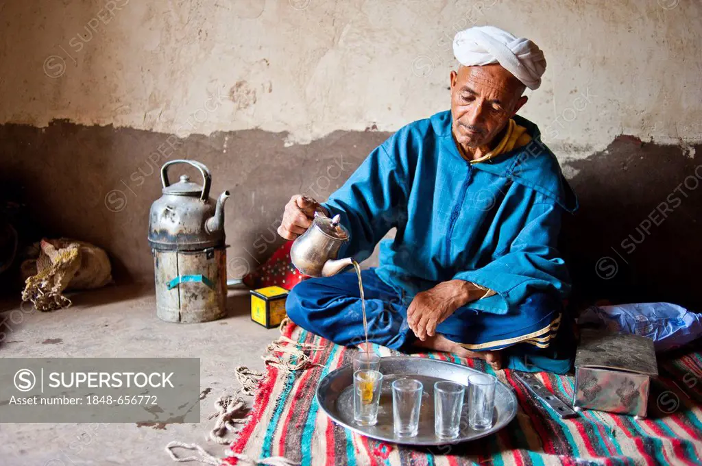 Elderly Berber man wearing a turban sitting on the floor on a rug pouring traditional mint tea from a silver jug, Kelaa M'gouna, High Atlas Mountains,...