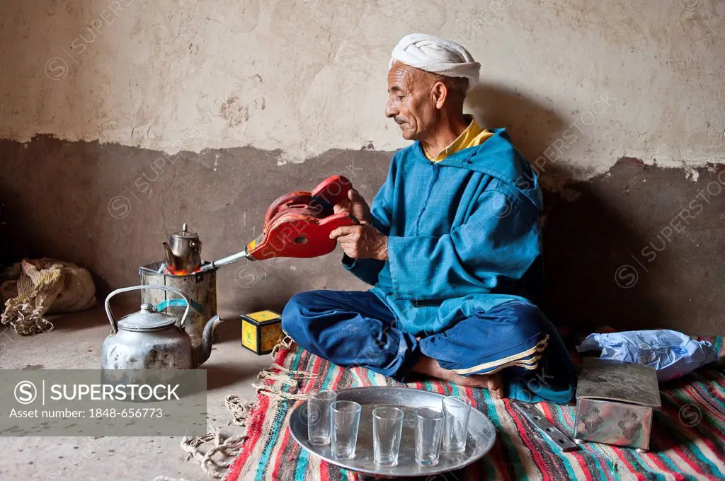 Elderly Berber man wearing a turban sitting on the floor on a rug preparing traditional mint tea, kindling the fire in a little tin stove with a blowe...