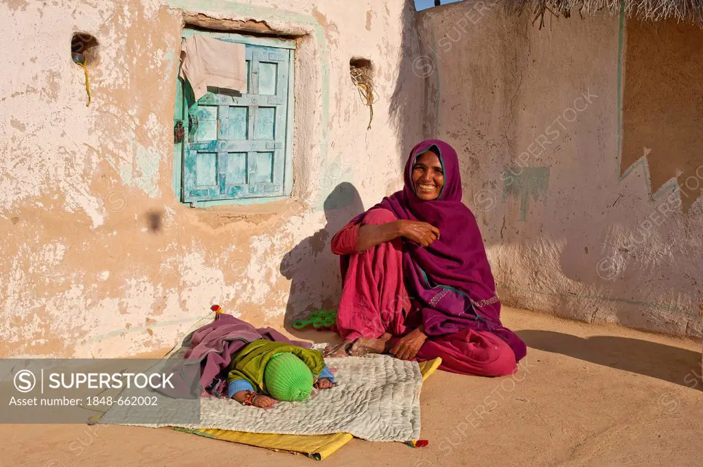 A friendly Indian woman wearing a red sari sitting on the floor in her courtyard, her sleeping child is lying in front of her on a blanket, Thar Deser...