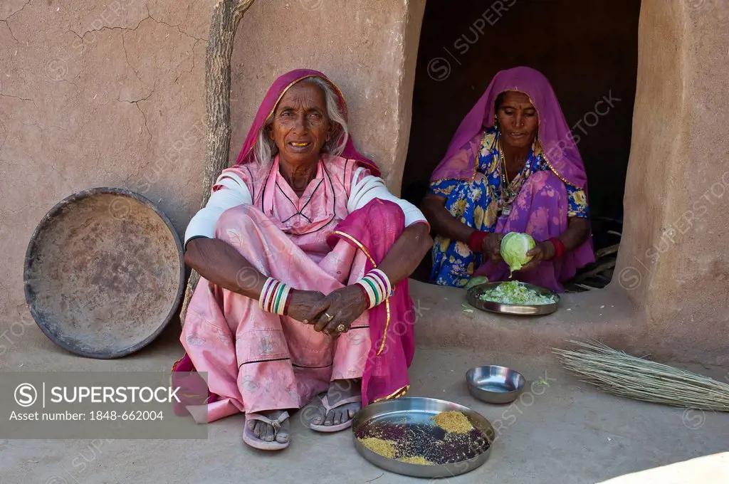 An elderly Indian woman wearing a sari sitting on the floor in front of a house entrance, a younger woman is sitting at the door and preparing a cabba...