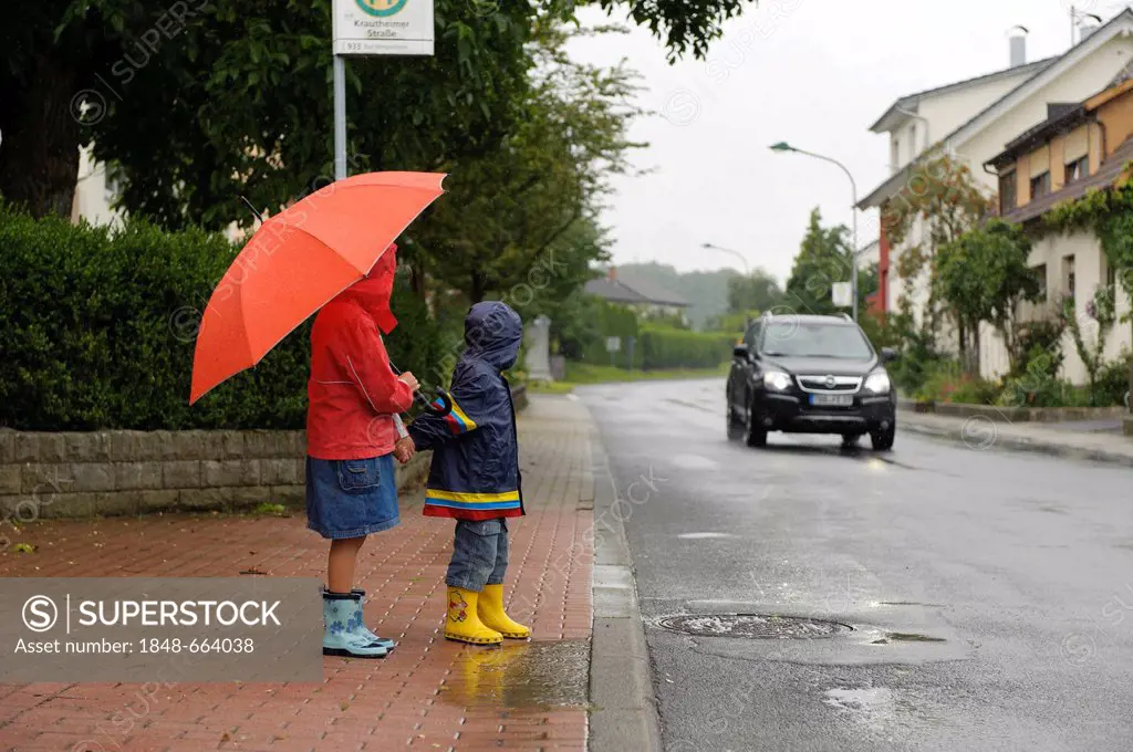 Two children, 3 and 7 years, waiting to cross the street in the rain while a car approaches, Assamstadt, Baden-Wuerttemberg, Germany, Europe