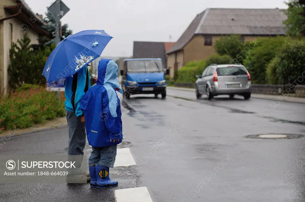Two children, 4 and 8 years, waiting to cross the street in the rain, two cars, Assamstadt, Baden-Wuerttemberg, Germany, Europe