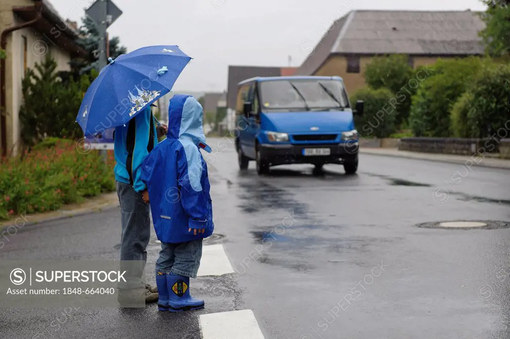 Two children, 4 and 8 years, waiting to cross the street in the rain while a car approaches, Assamstadt, Baden-Wuerttemberg, Germany, Europe