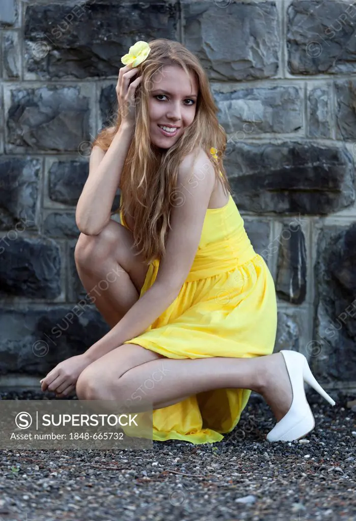 Young woman in a yellow dress and a yellow flower posing in a crouching position in front of a stone wall
