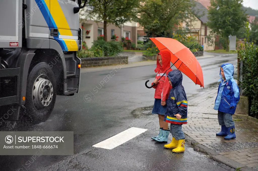 3 children, 3 to 7 years, crossing the main road in the rain, Assamstadt, Baden-Wuerttemberg, Germany, Europe