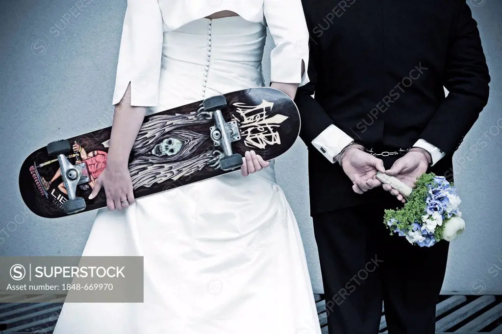 Wedding couple seen from behind, the bride is holding a skateboard, the groom is wearing handcuffs and holding a bridal bouquet