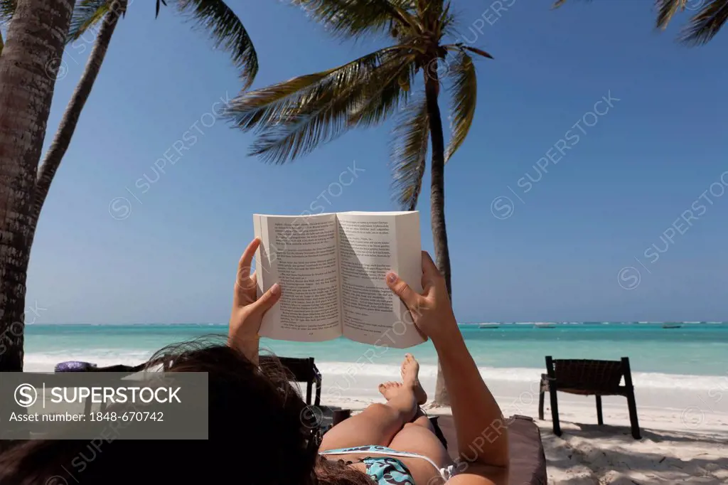 Girl, 16 years, reading a book on the beach under palm trees