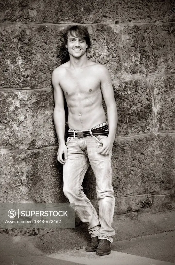 Bare-chested young man wearing jeans, smiling, leaning against an old wall