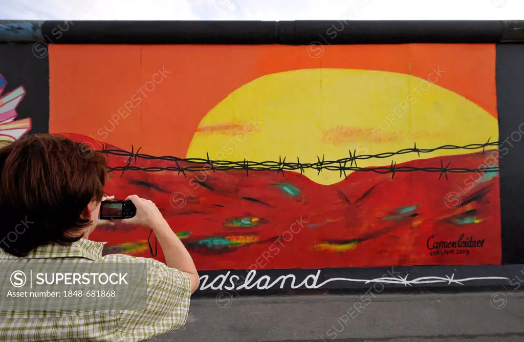 Tourist taking a photograph of Art and the Berlin Wall, sunrise in the East behind barbed wire, painting on a remaining segment of the Berlin Wall, Ea...