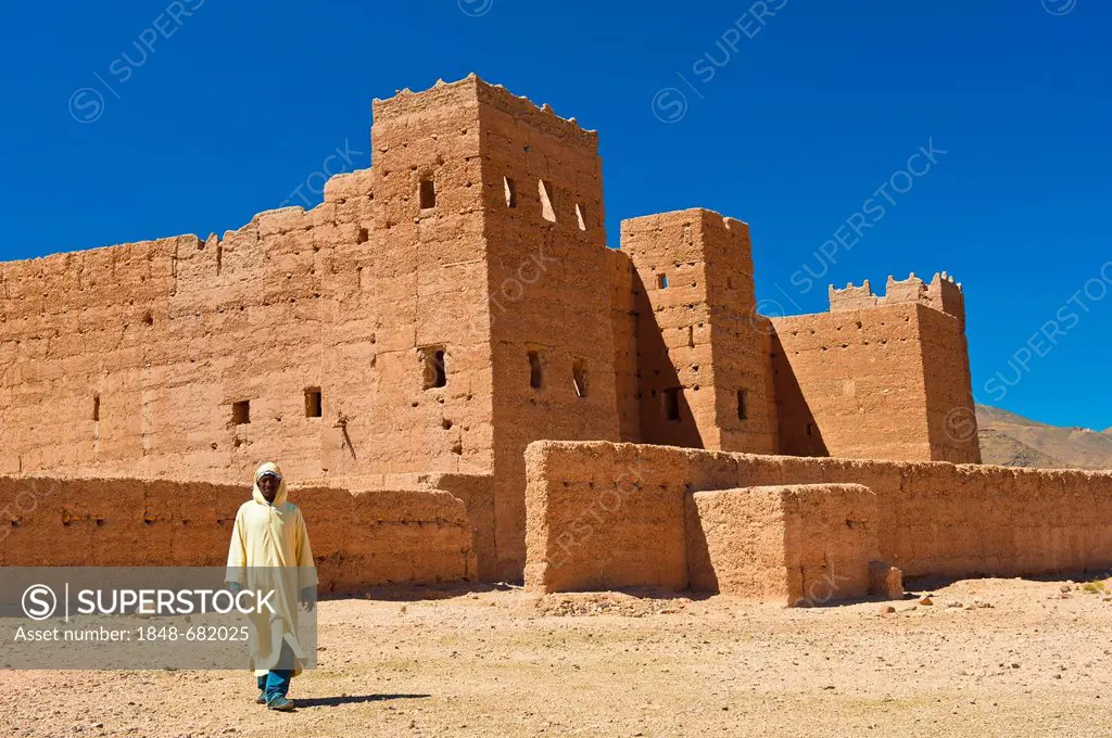 Berber man wearing a traditional djellabah walking in front of Taouirt Kasbah, mud fortress, mud brick building of the Berber tribes, Tighremt, Draa V...
