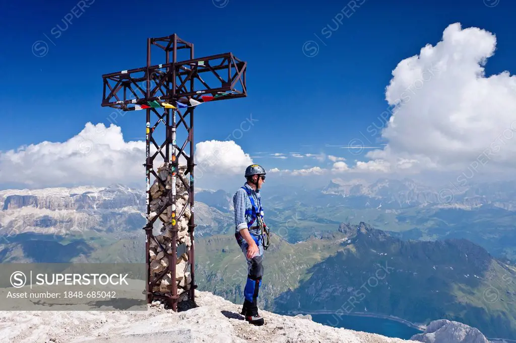 Mountaineer standing next to the summit cross of Marmolada mountain, Dolomites, Passo Fedaia mountain pass, Heiligkreuzkofel group and Sella group at ...