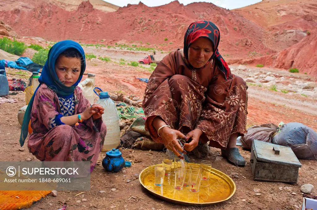 Cave dwelling nomads, a woman and a little girl sitting in front of a cave dwelling, the woman is cleaning tea glasses on a brass tray, High Atlas Mou...