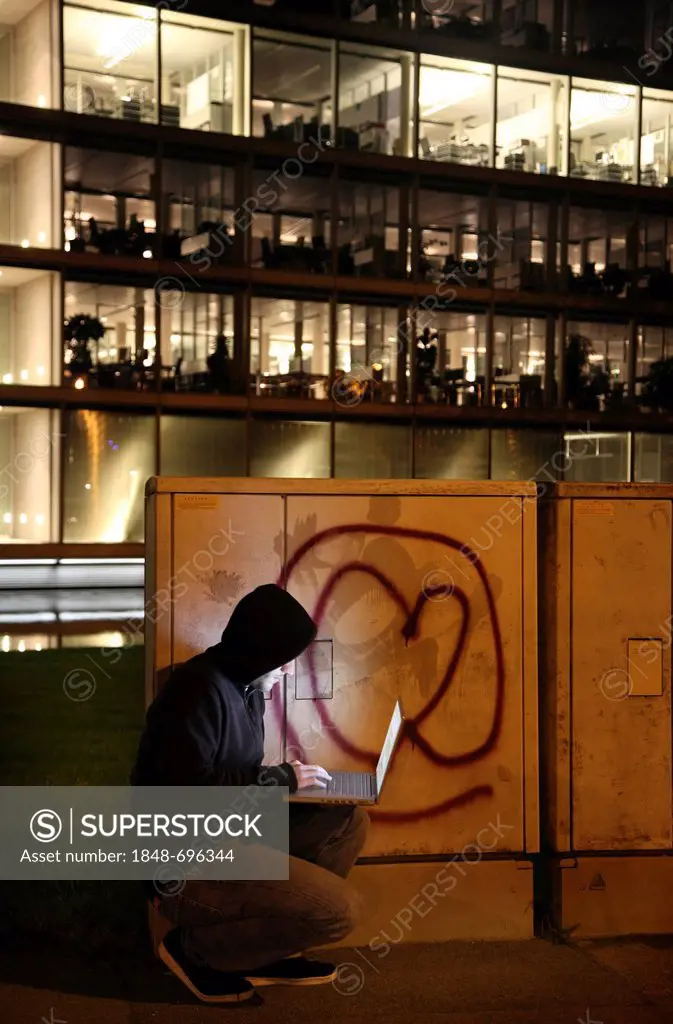 Hacker working on a laptop computer in front of a commercial building, hacking into the company's network, symbolic image for computer hacking, comput...