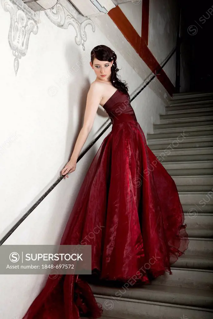 Young woman wearing a red dress with a long train standing on the stone stairs in a stairwell