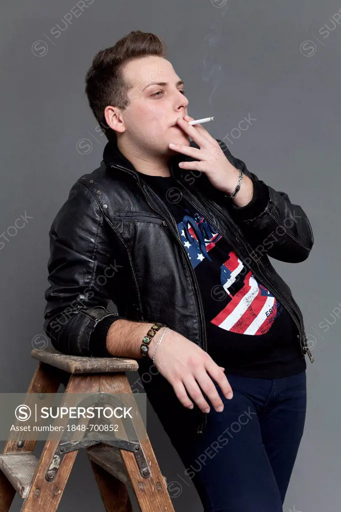 Young man smoking a cigarette wearing a leather jacket and jeans while leaning casually against an old wooden ladder