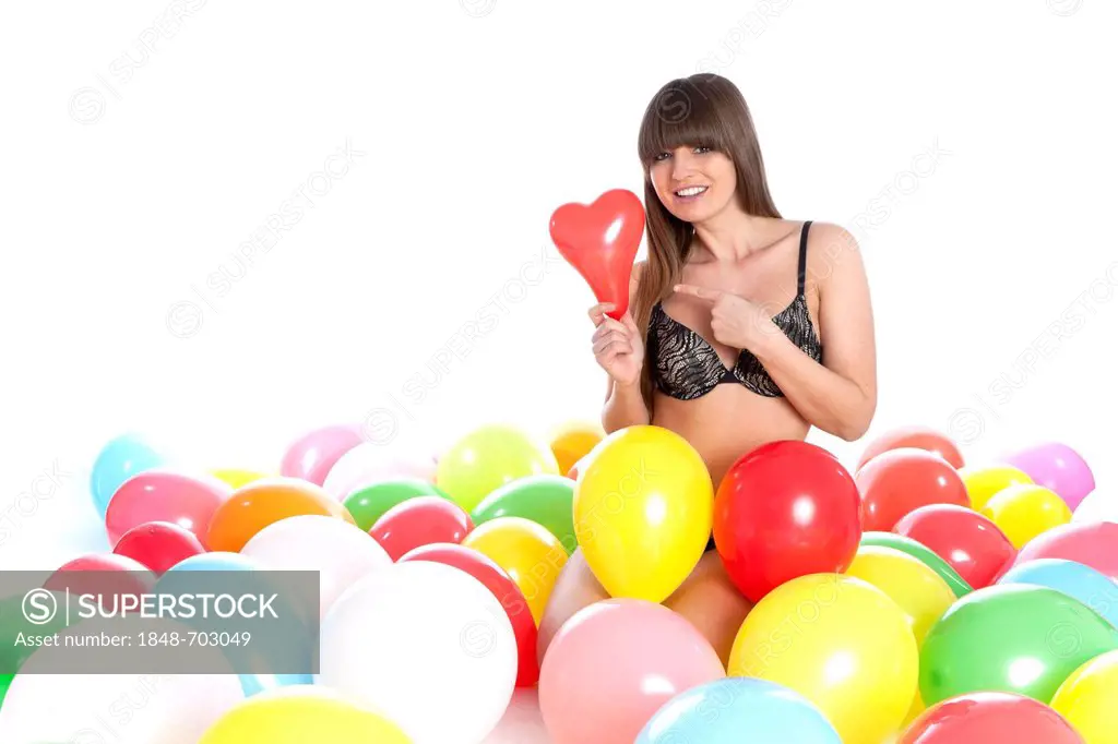 Young woman wearing lingerie sitting between colourful balloons, holding and pointing to a heart-shaped balloon