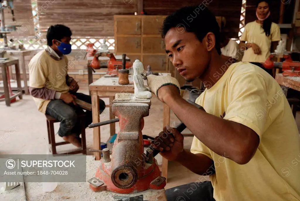 Two young Cambodian artisans during the artistic craftsmanship of stone with a wooden hammer and a chisel to produce a traditional Buddha figure, Arti...