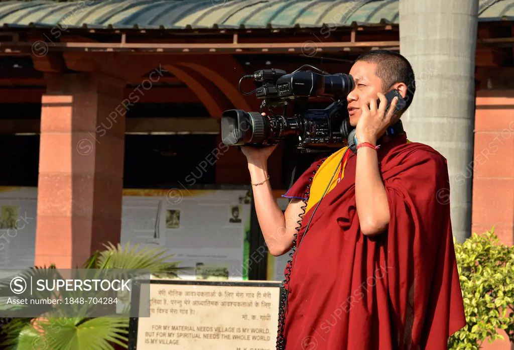 Tibetan monk in a red robe working as cameraman carrying a large video camera and a mobile phone, Global Buddhist Congregation 2011, at Smitri Gandhi,...