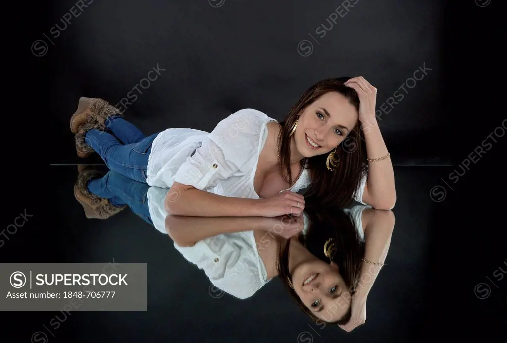 Young smiling woman wearing a white top and jeans, lying on a mirror, mirrored image
