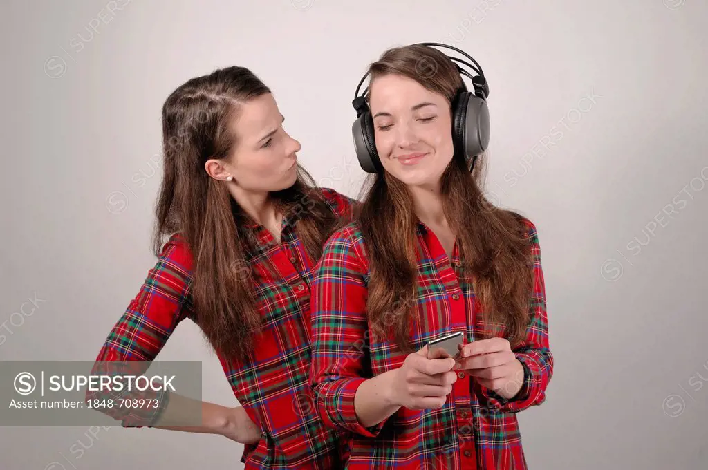 Twin sisters, one holding an iPod and listening to headphones, the other looking enviously