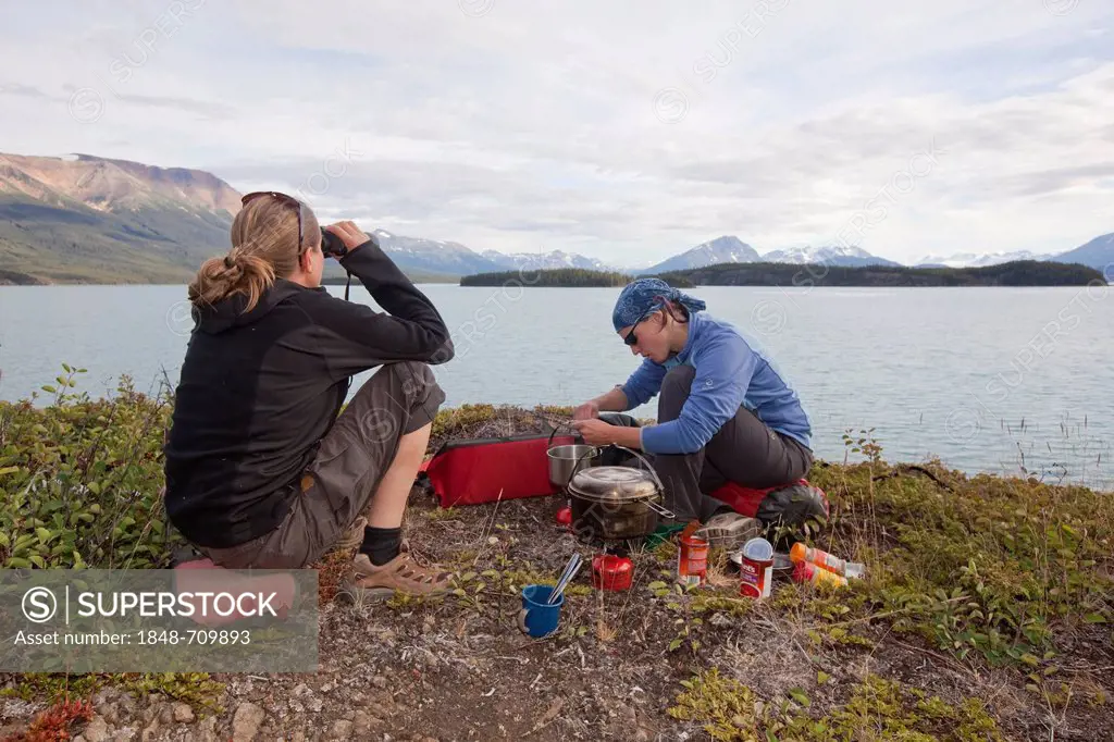Two young women cooking on a camping stove, watching lake with binoculars, Atlin Lake, British Columbia, Canada, America