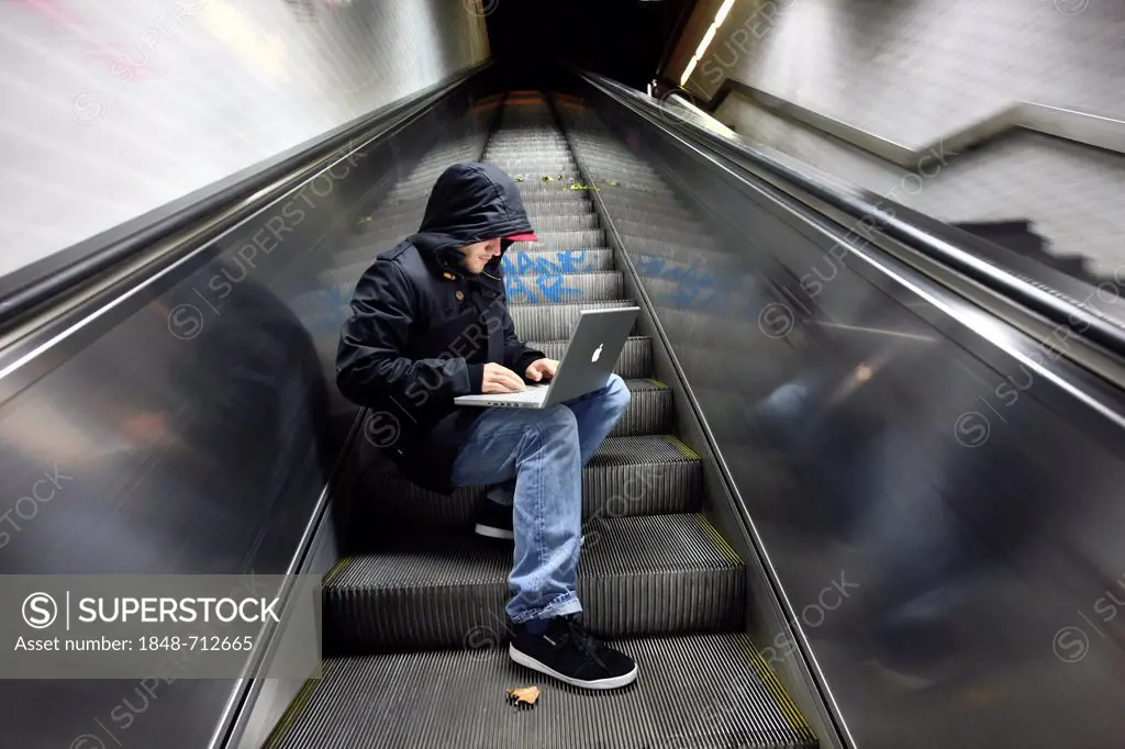 Hacker working on a laptop computer on an escalator in a subway passage at night, symbolic image for computer hacking, computer crime, cybercrime, dat...