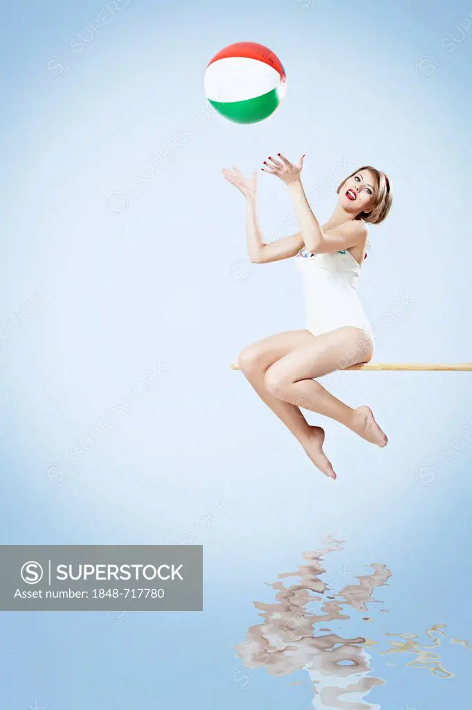 Young woman wearing a white bathing suit sitting on a diving board and trying to catch a beach ball, pin-up