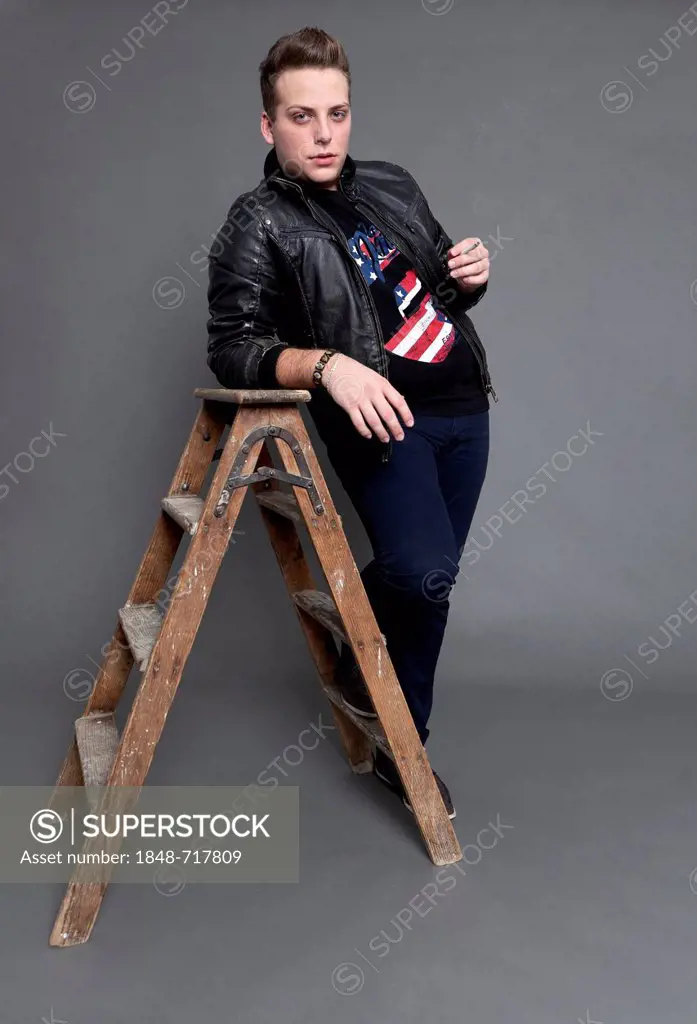 Young man with a cigarette wearing a leather jacket and jeans while leaning casually against an old wooden ladder
