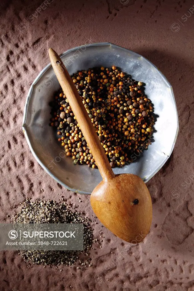 Colourful peppercorns (Piper nigrum) on a metal plate with a wooden spoon and coarsely ground pepper on a rustic stone base