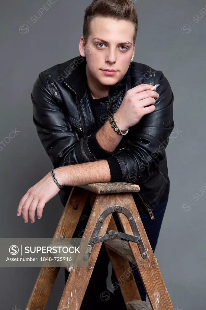 Young man in a leather jacket smoking a cigarette, leaning on a ladder