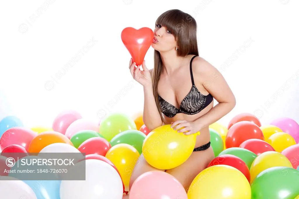 Young woman in lingerie sitting amidst balloons, kissing red balloon heart
