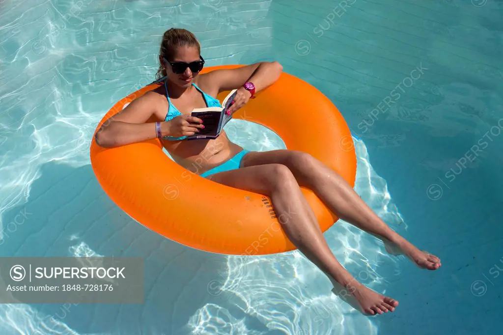 Young woman sitting on an inner tube, floating tyre, reading a paperback in a private swimming pool, Germany
