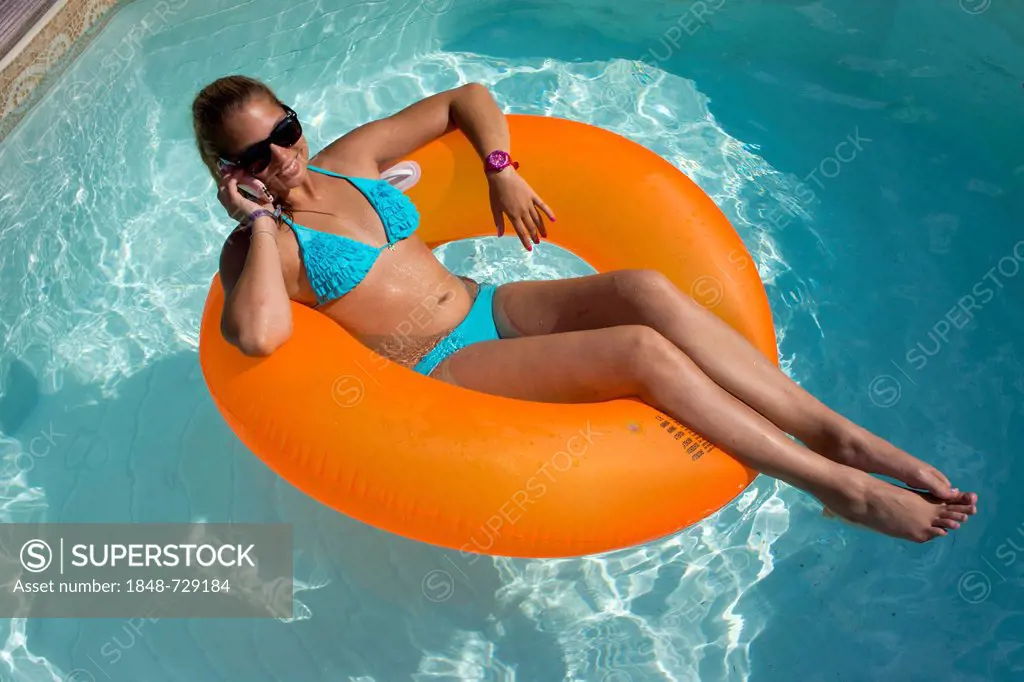 Young woman sitting on an inner tube, floating tyre, talking on a mobile phone in a private swimming pool, Germany