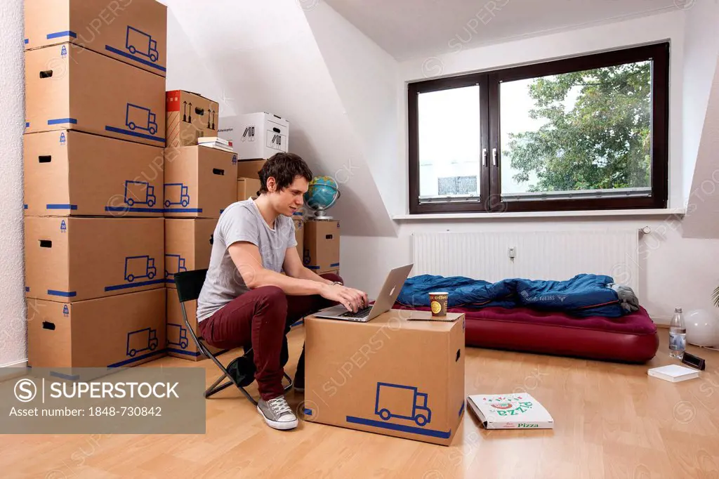 Young man sitting in an empty room in a new apartment, provisionally furnished with a sleeping bag, an air mattress, a laptop and moving boxes