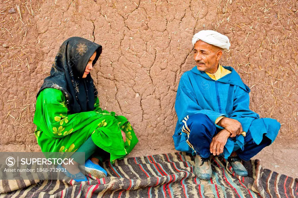 Elderly man and a young girl, Berber people, squatting on a carpet or rug in front of a mud brick house talking, High Atlas mountain range, Morocco, A...