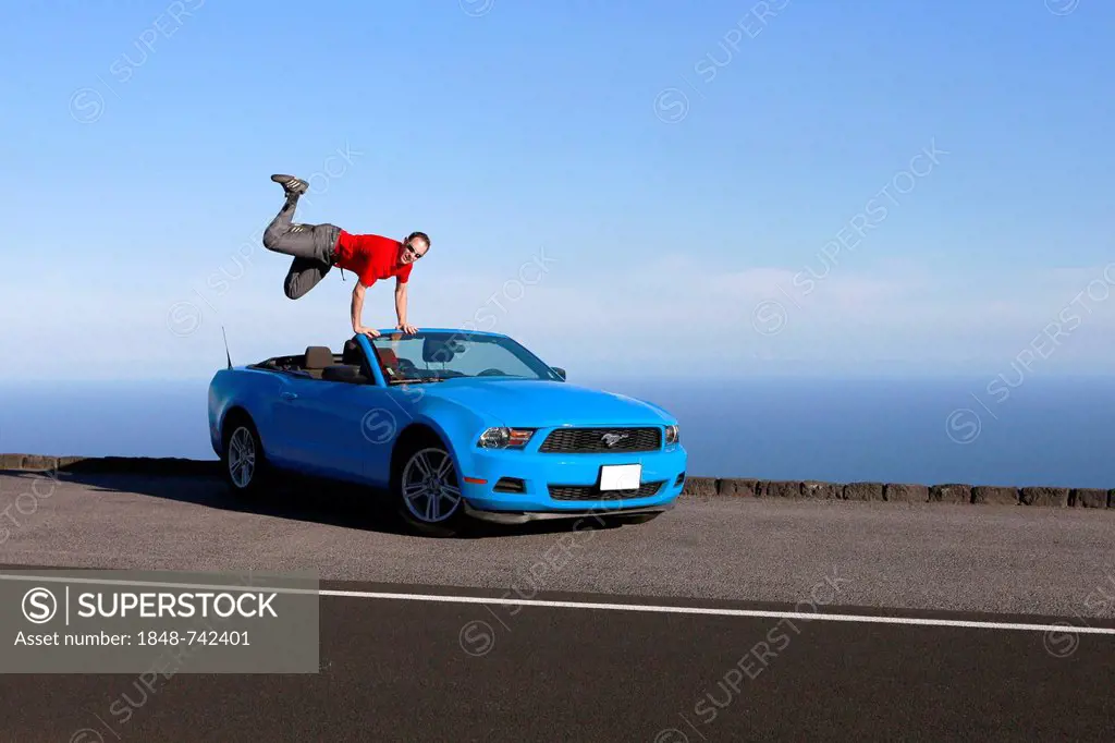 Man jumping with joy on a sky-blue Ford Mustang convertible by the sea, Big Island, Hawaii, USA