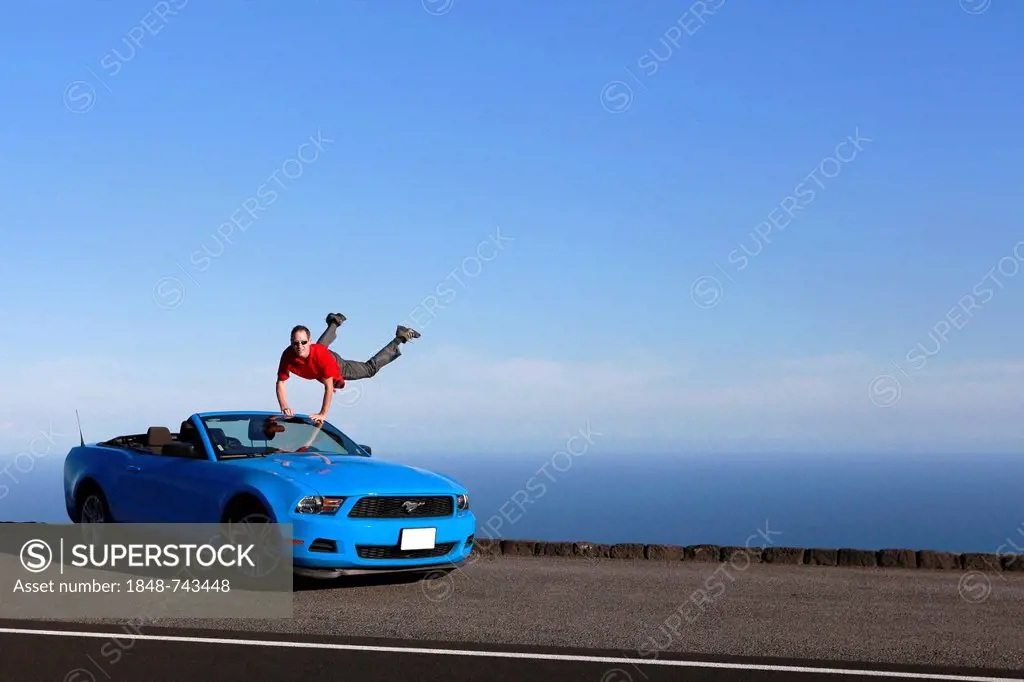 Man jumping with joy on a sky-blue Ford Mustang convertible by the sea, Big Island, Hawaii, USA