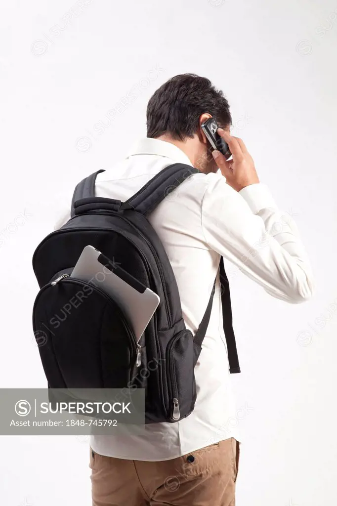 Man wearing a backpack, from which an iPad is sticking out, talking on a mobile phone