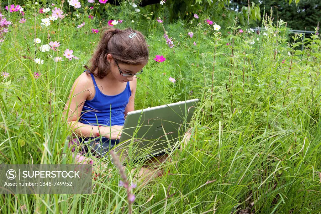 Girl, 11 years, sitting on a meadow working on a laptop computer