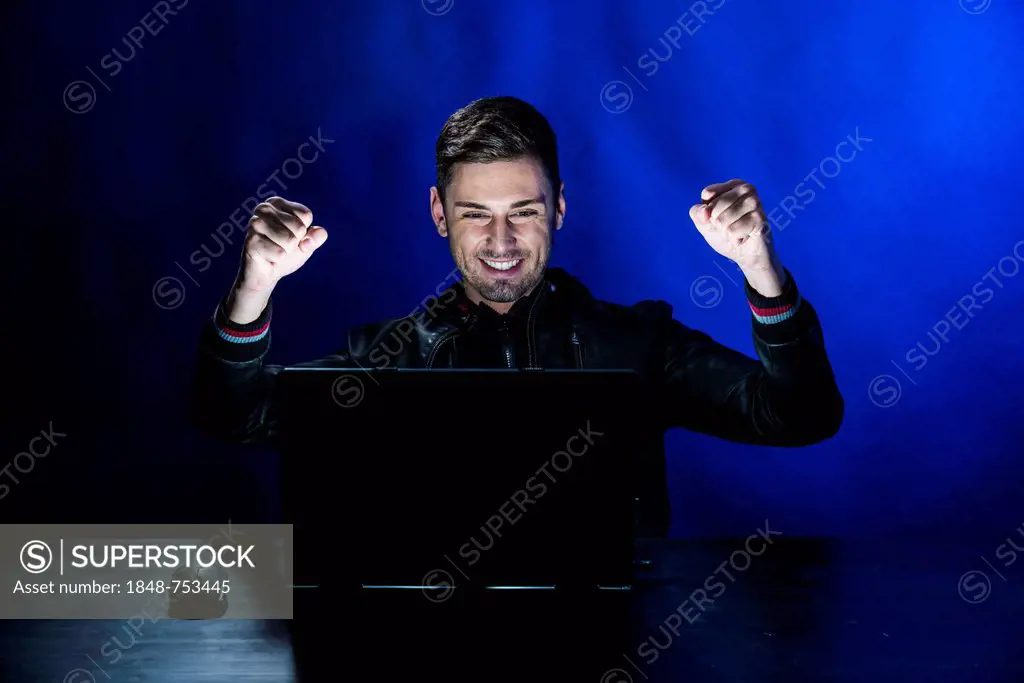 Young man sitting in the dark in front of a laptop, with a joyful expression, exulting